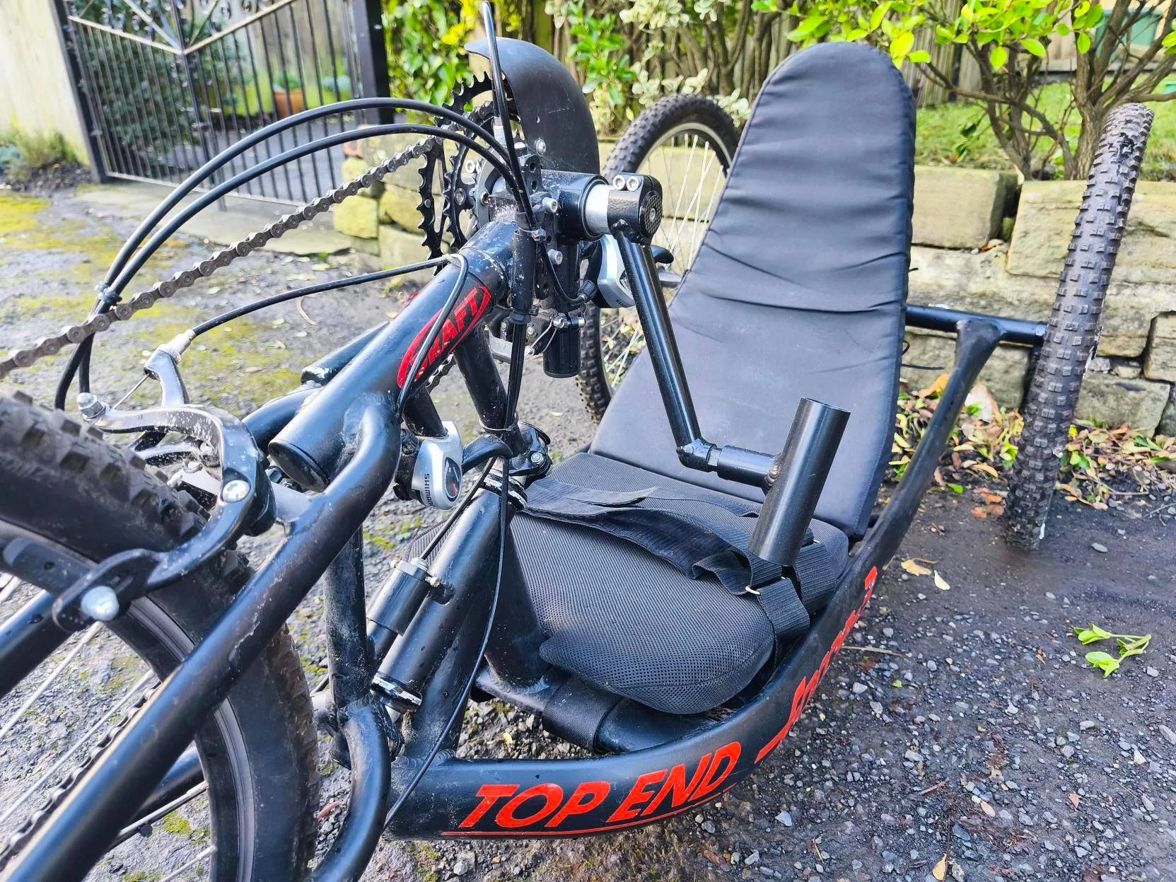 Accessible black handcycle with padded seat on a wet path by a garden, supporting disabled access to outdoor community activities.