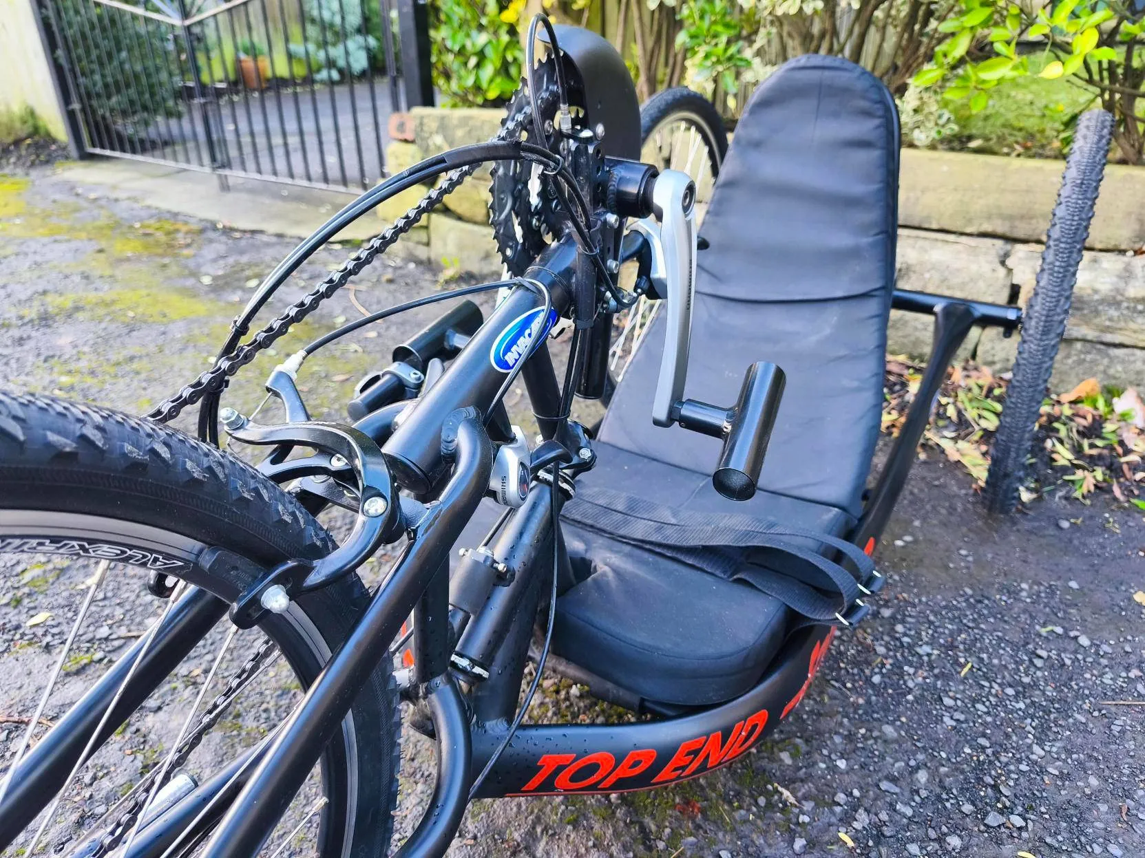 Black TOP END handcycle with padded seat on gravel drive near plants, promoting disabled access to outdoor activities and green spaces.