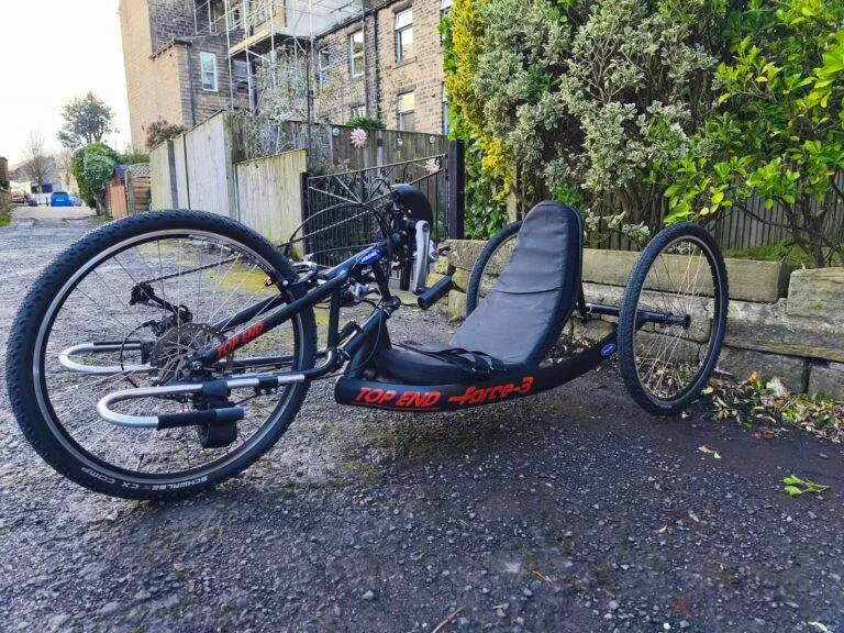 Accessible black Top End Force-3 handcycle by green bushes on a paved path, supporting disabled outdoor activities and cycling.